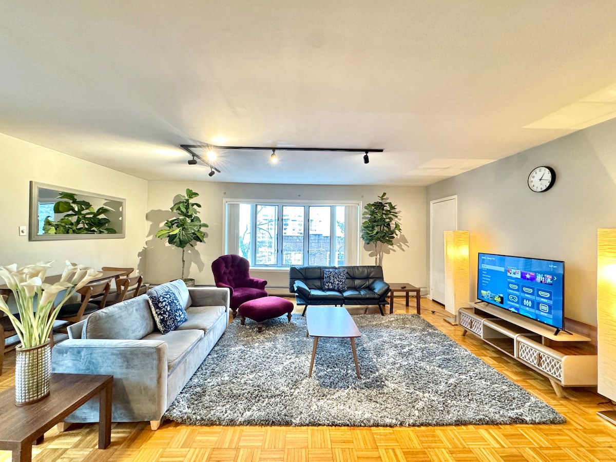 The spacious living area features a comfortable arrangement of seating, including a grey sofa, black couch, and a purple accent chair. A large window allows natural light to fill the room, while a television occupies a modern entertainment unit. Decorative plants add a touch of greenery.