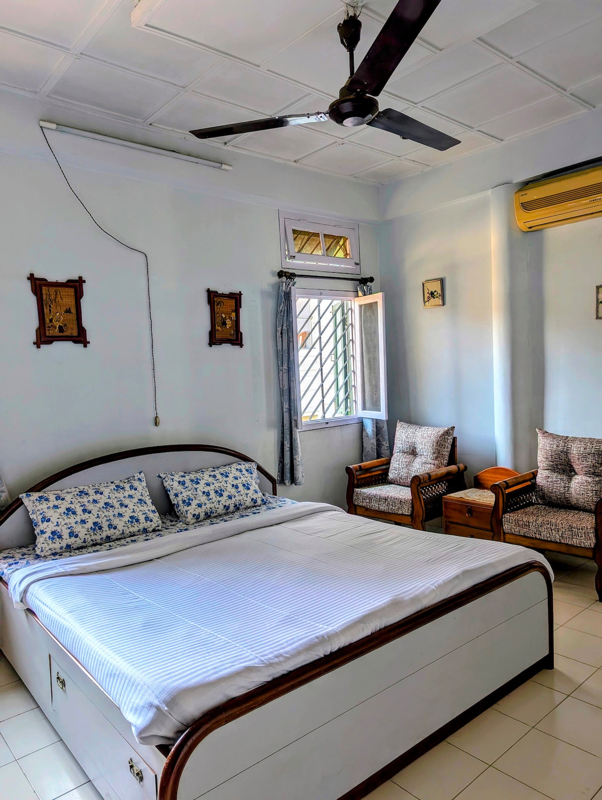 A well-lit sleeping area features a spacious bed with a simple white bedspread and floral accent pillows. Two cozy armchairs are positioned nearby, complemented by wooden furniture. A window allows natural light to illuminate the room, enhancing the calming ambiance.