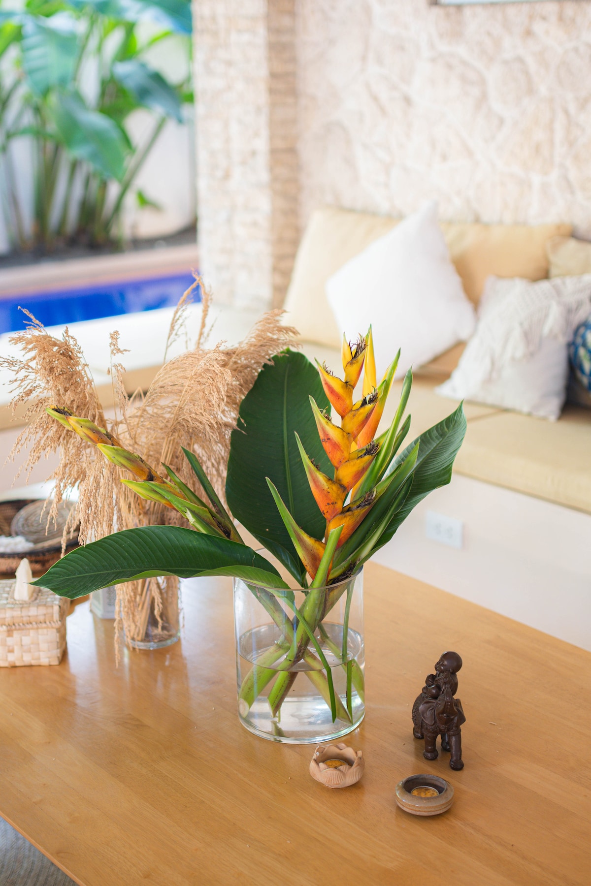 A coffee table displays a glass vase filled with vibrant orange and green tropical flowers. Dried ornamental grasses complement the arrangement. Surrounding decor includes a small wooden figurine and a round dish, set against a backdrop of a light-colored couch and a glimpse of a pool outside.