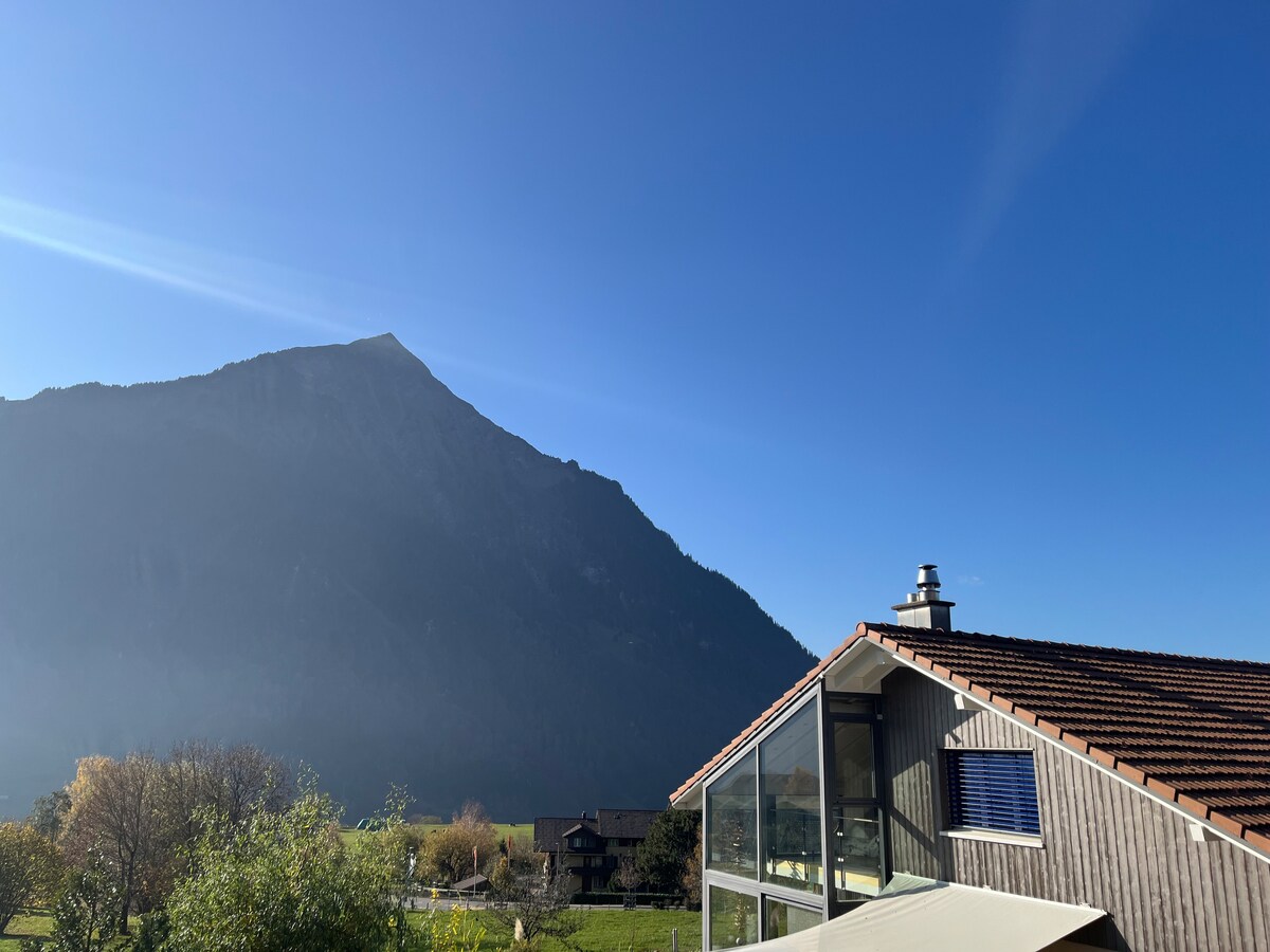 A mountain rises prominently against a clear blue sky, providing a striking backdrop. The image captures a portion of a modern building with large glass windows reflecting the natural surroundings. Lush greenery is visible in the foreground, enhancing the serene setting.