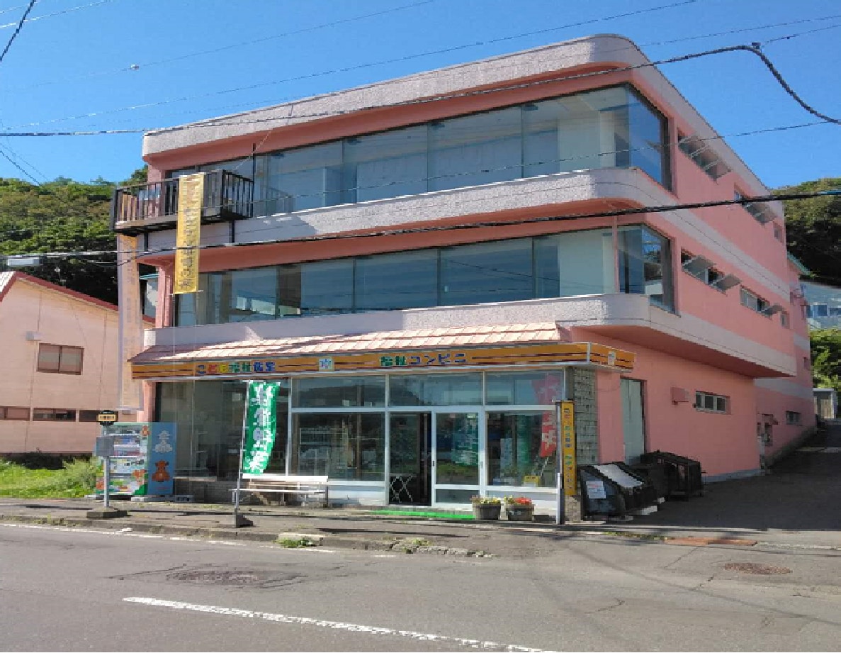 A three-story pink building is presented, featuring large glass windows and a welcoming entrance on the ground floor. A balcony is visible on the upper level. Surrounding foliage adds a natural touch to the urban setting.