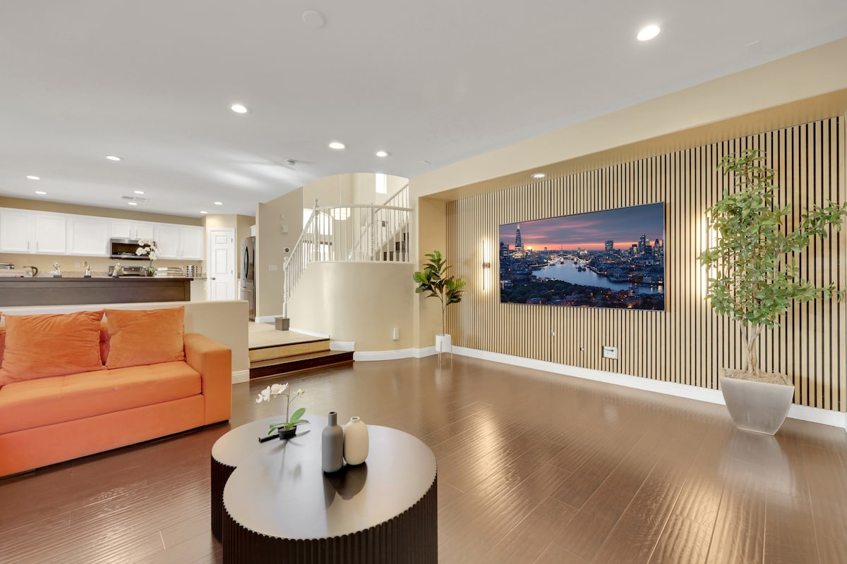 A modern living area features an orange sofa and a round coffee table with decorative items. A flat-screen TV is mounted on a wooden accent wall. Recessed lighting highlights the spaces, while a staircase and dining area are visible in the background.