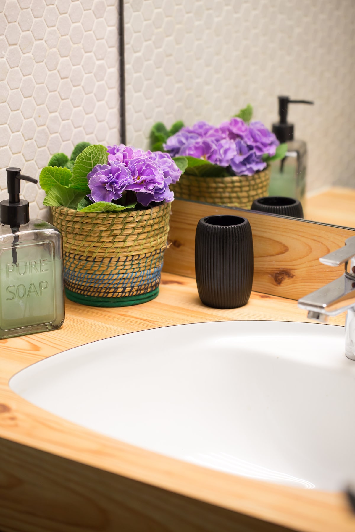 A bathroom sink area features a white basin set against a wooden countertop. A woven basket holds vibrant purple flowers, accompanied by a glass soap dispenser labeled 'PURE SOAP' and a sleek black vase. Reflections in the mirror enhance the overall lightness of the space.