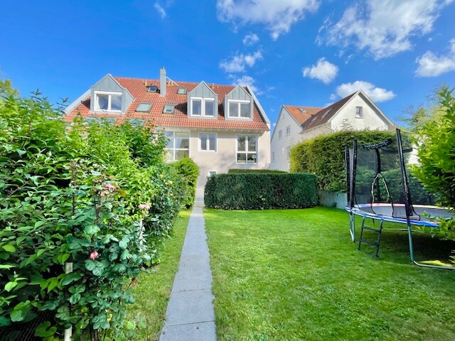 A spacious garden area is bordered by well-maintained hedges, leading to a modern building with a red-tiled roof. A grassy lawn features a trampoline, and the sky above is bright with scattered clouds.
