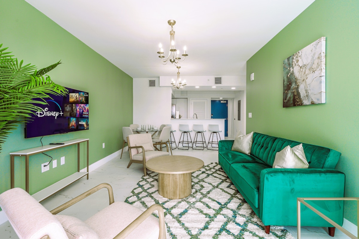 A modern living area features green walls, a chic emerald sofa, and a round coffee table atop a textured rug. A wall-mounted TV displays the Disney+ logo. A well-lit dining area is visible in the background, with a stylish kitchen space beyond.