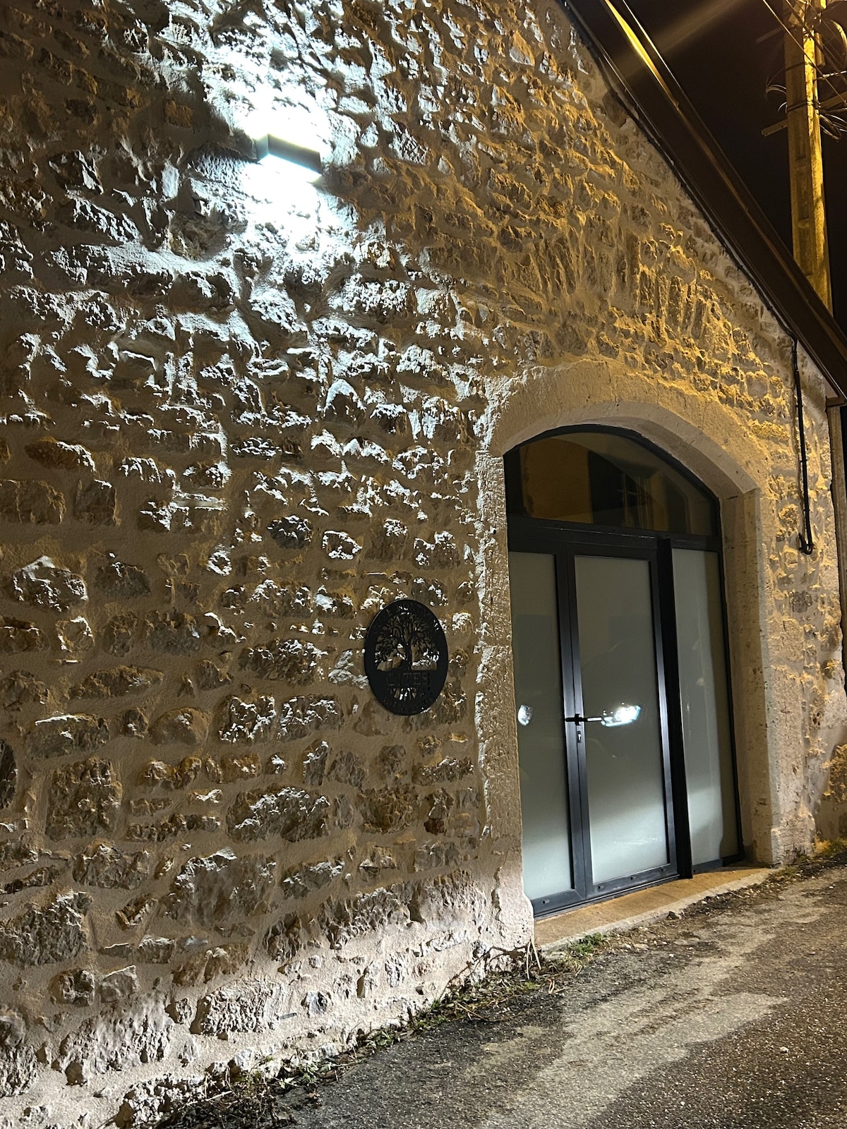 The exterior of a stone building is illuminated at night, showcasing its textured surface. A large glass door is framed by an elegant archway, while a circular wall sign is affixed beside the entrance. Soft lighting highlights the building's features.