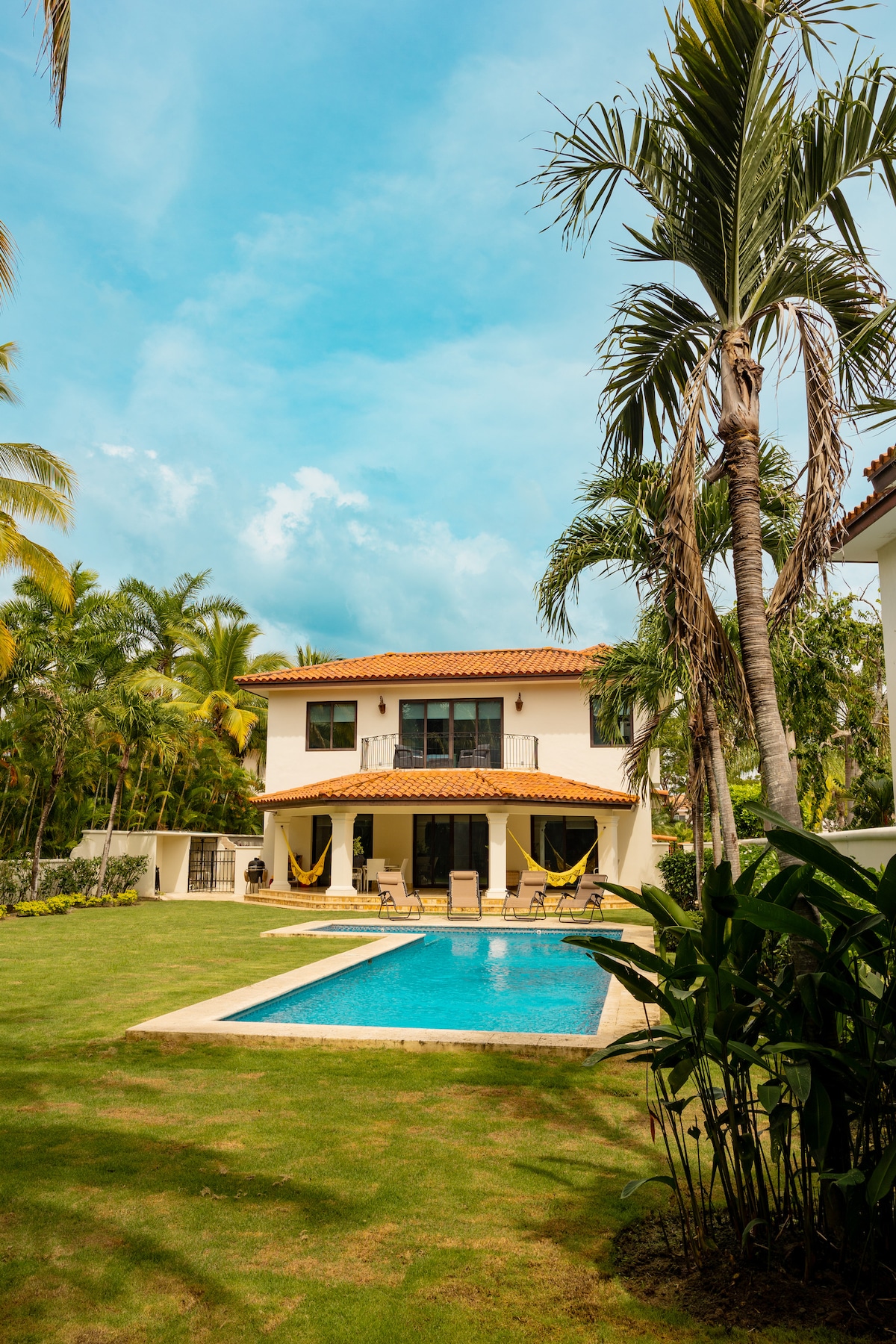 The exterior of the house features a spacious lawn surrounded by lush palm trees. A private pool is centrally located, visible in shades of blue. The two-story structure showcases a warm-coloured roof and large windows, complemented by hanging hammocks on the terrace.