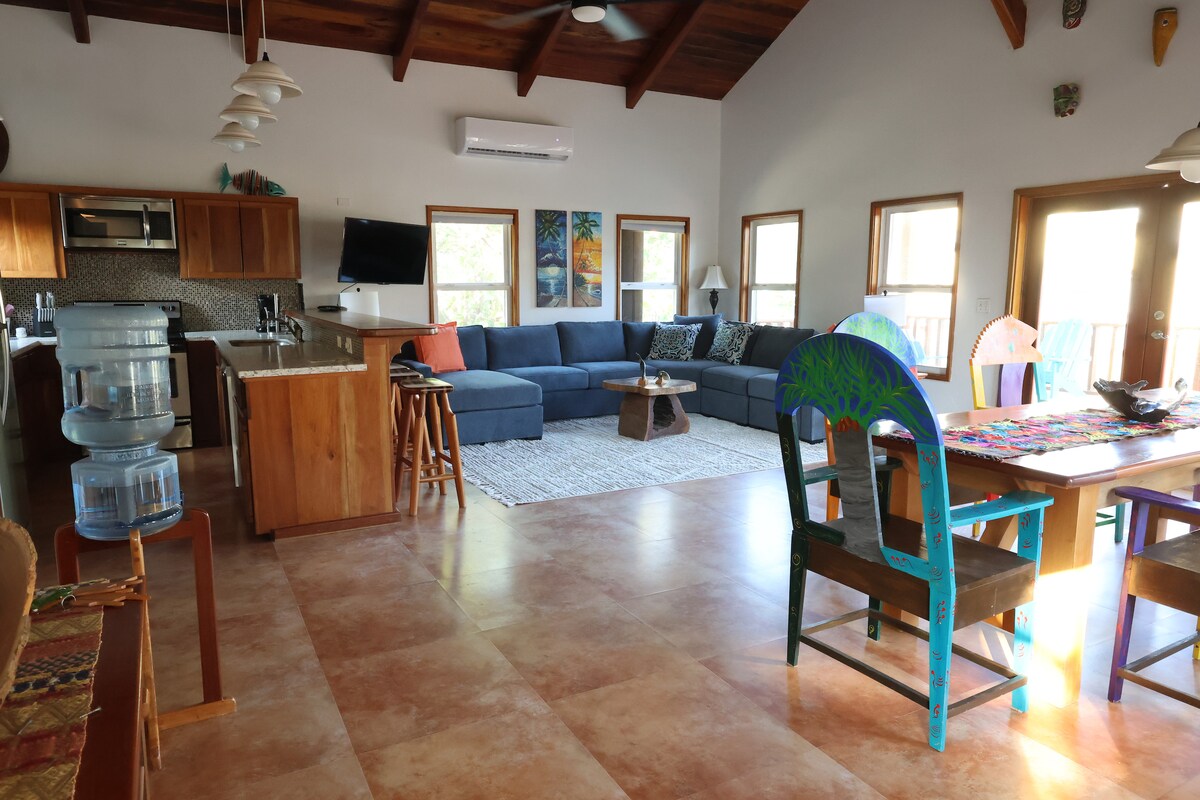 An open living space features a sectional sofa in a muted blue tone, paired with a rustic coffee table and colorful dining chairs. A large window allows natural light to fill the area, highlighting the wooden beams of the ceiling and the adjacent kitchen.