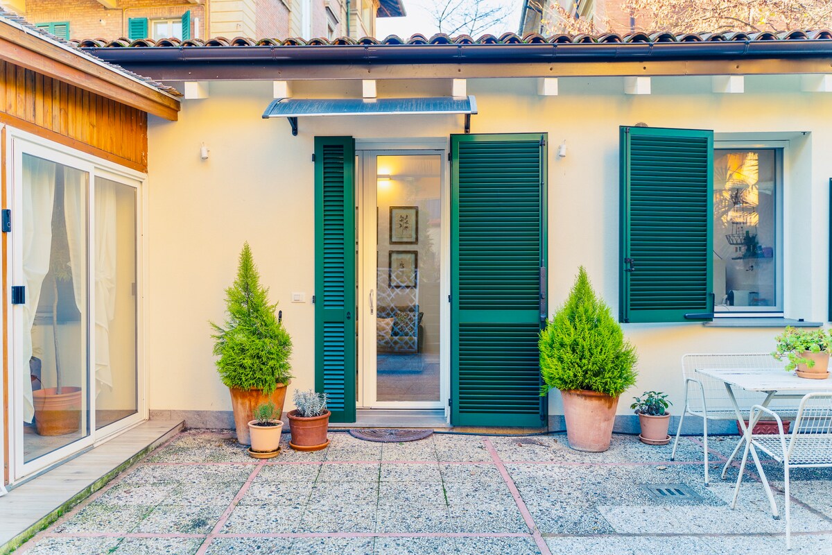The entrance features light-colored walls with green shutters on either side of a glass door. Potted plants are arranged on each side of the doorway. A small outdoor dining area is visible, with a white table and chairs set on a stone patio.