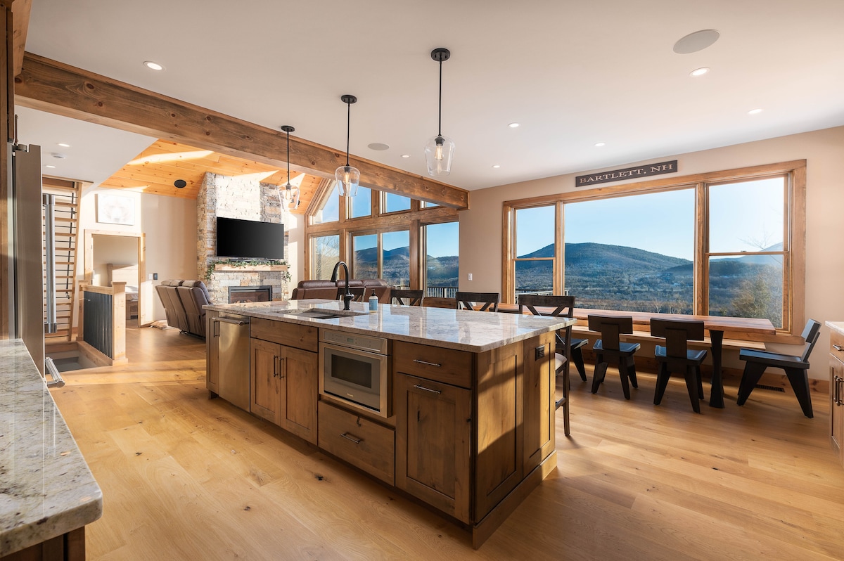 A spacious kitchen features a large island with modern appliances and a built-in microwave. Expansive windows offer views of the mountains, while the dining area with a long table is visible in the background, complementing the warm wood accents and natural light.