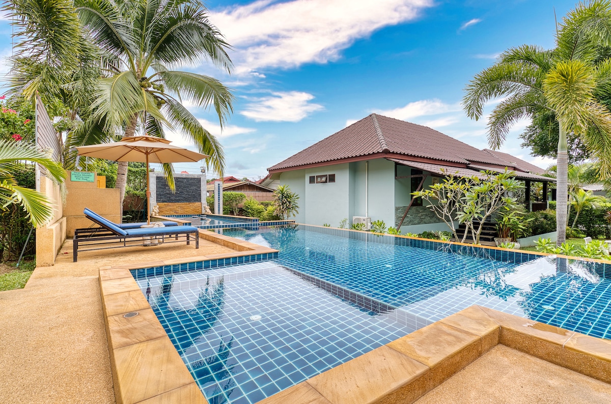 A serene shared pool is surrounded by a well-maintained tropical garden. Comfortable lounge chairs are positioned under an umbrella, with palm trees and colorful foliage creating a relaxing environment. The house is visible in the background, contributing to the overall tranquil setting.