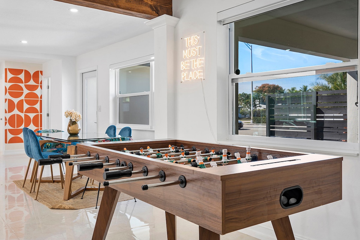 A modern living area features a foosball table in the foreground, surrounded by open, bright spaces. In the background, a dining area with stylish chairs and a large window allows for plenty of natural light, enhancing the inviting atmosphere.