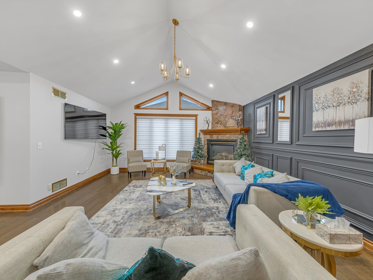 A spacious living area is highlighted by a vaulted ceiling with modern light fixtures. Soft furnishings include a light-colored sofa and armchairs, arranged around a coffee table. A fireplace serves as a focal point, flanked by decorative plants and artful wall panels.