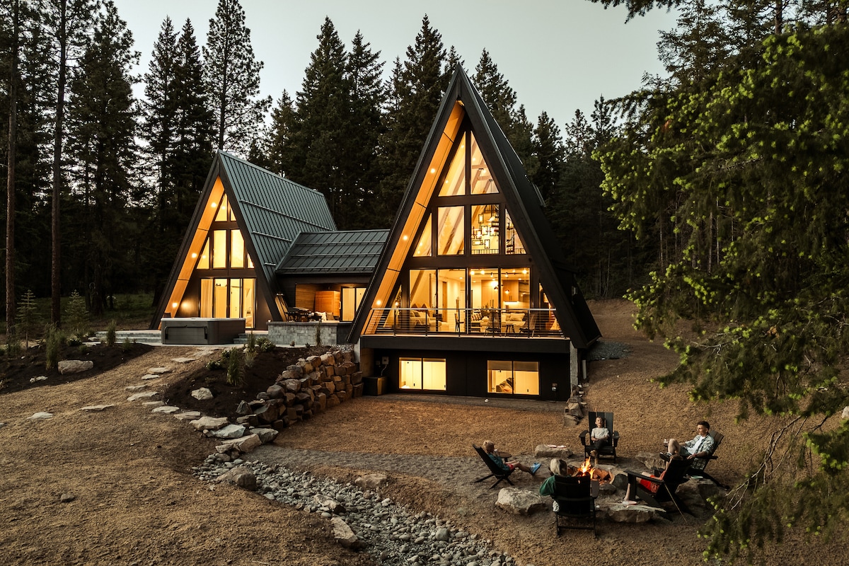 The double A-frame structure is surrounded by towering trees and features expansive glass windows illuminated from within. A cozy outdoor fire pit area is positioned in the foreground, along with seating arranged around it. A pathway lined with stones leads up to the entrance.