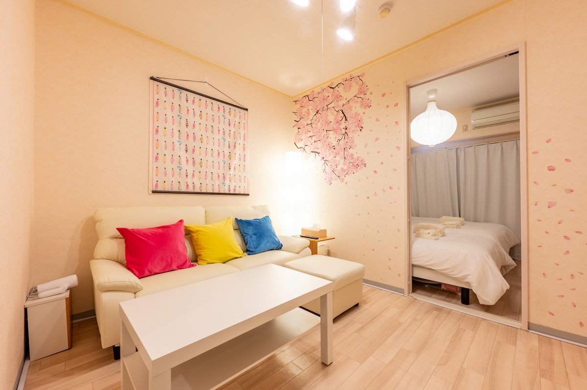 A living area features a light-colored sofa adorned with colorful cushions. A white coffee table sits in front, with a soft glow from nearby lighting. A door leads to a separate bedroom, and pink cherry blossom wall art adds a charming touch.