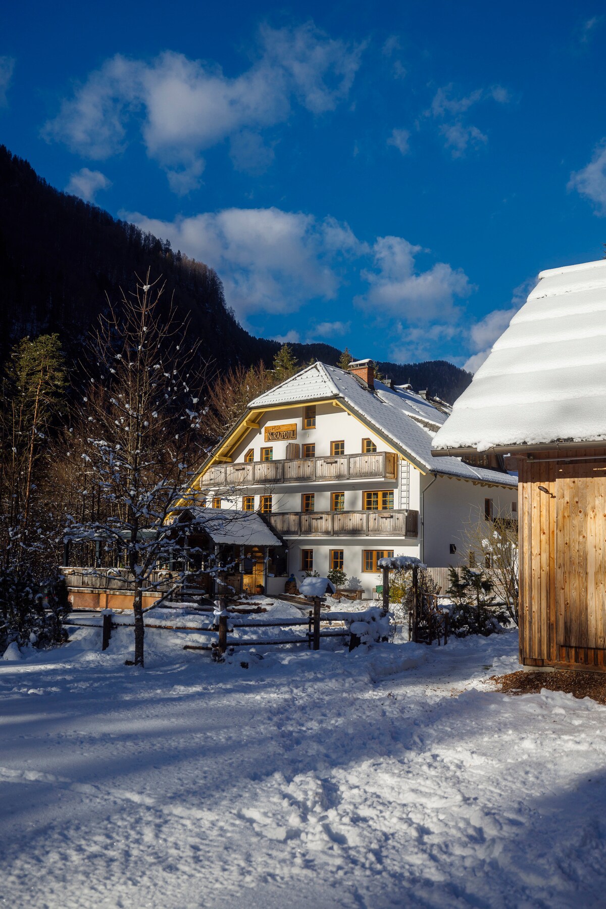 The exterior of a multi-level building is shown, featuring a white facade and wooden accents. Balconies extend from the upper levels, surrounded by snow-covered grounds and tall trees. A nearby wooden structure complements the scene, set against a backdrop of mountains and a bright blue sky.