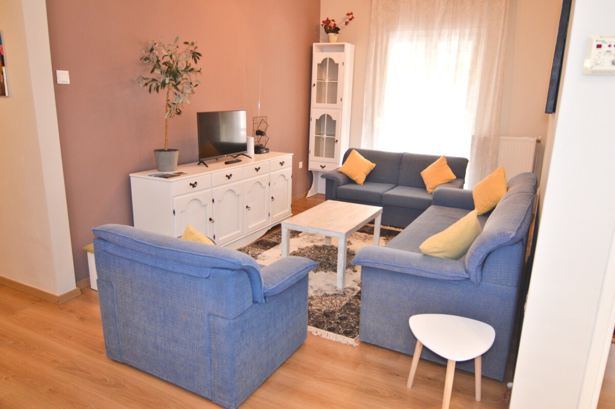 A living area is presented with a white television stand and a flat-screen TV. Two blue sofas, adorned with yellow cushions, frame a light-colored coffee table on a decorative rug. A small side table complements the seating arrangement while natural light filters through the sheer curtains.