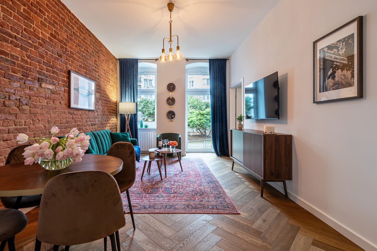 A living area showcases a mix of modern and vintage styles, featuring a brick accent wall and a plush teal couch. A round dining table with four chairs is positioned nearby. Soft lighting is provided by a chandelier, with a large window offering views of greenery.