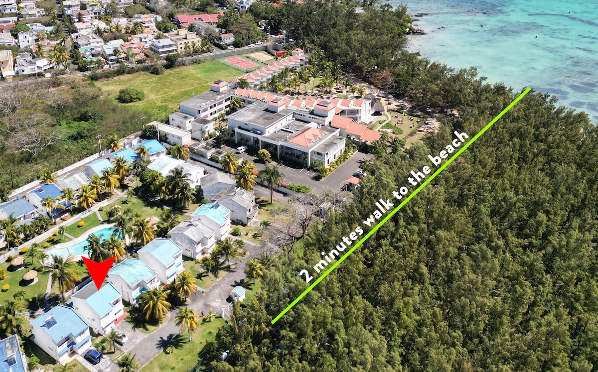 An aerial view captures the resort area with lush greenery and palm trees. The villa's location is highlighted, showing proximity to the beach, just a 2-minute walk away. The sparkling pool and outdoor spaces are visible within the landscaped complex.