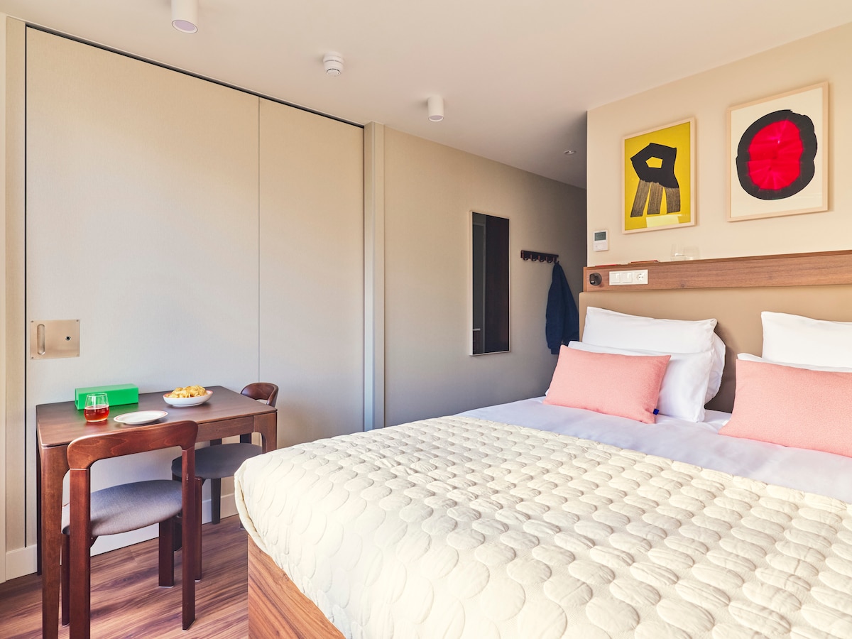 A modern studio features a spacious double bed dressed in light bedding and two decorative pillows. A small dining table, accompanied by two chairs, offers a cozy seating area. Wall art adds a touch of color to the neutral decor, enhancing the inviting ambiance.
