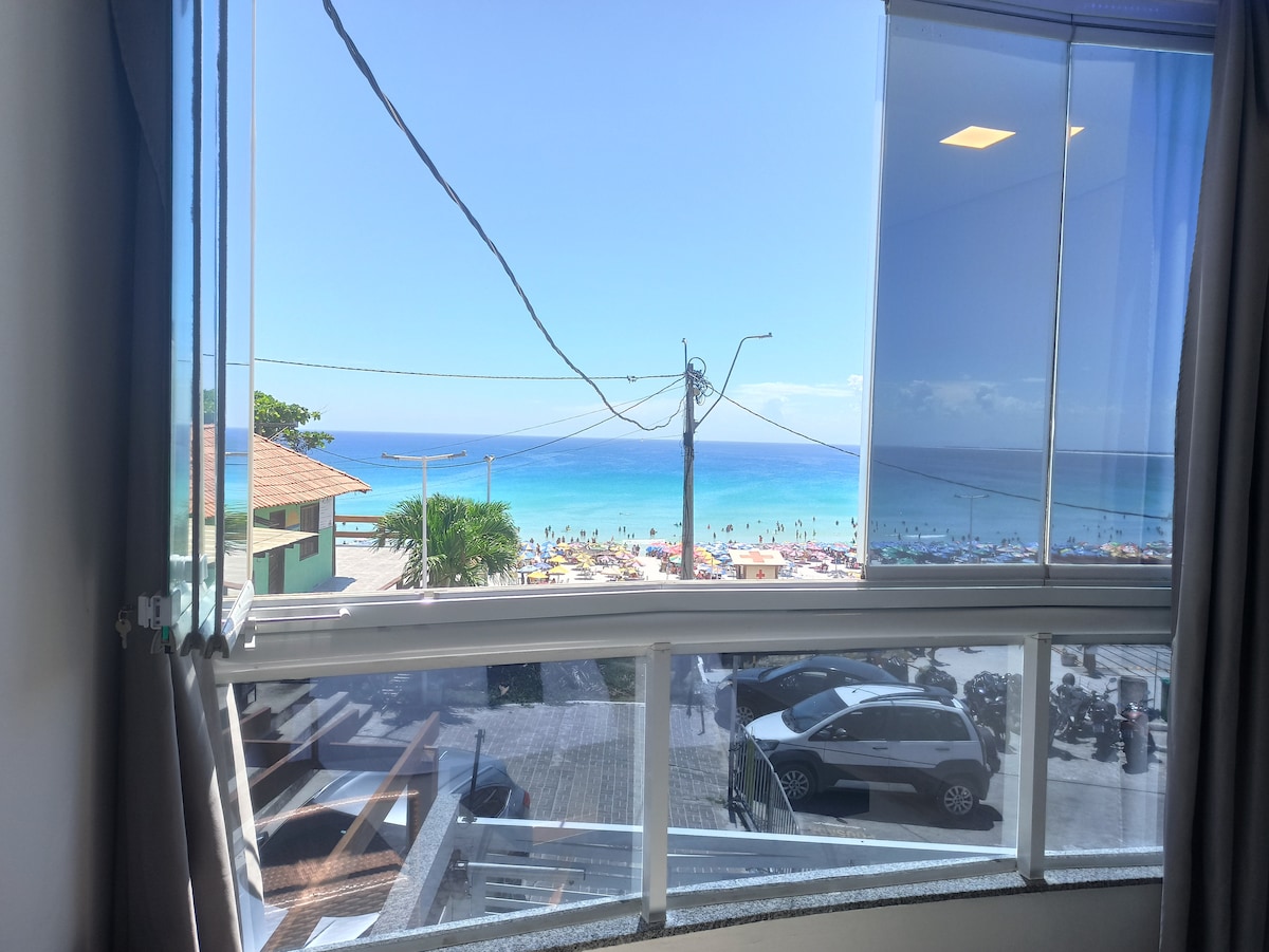 A panoramic view of the ocean is visible from the window, with the beach and colorful umbrellas dotted along the shore. The clear blue sky enhances the scene, while nearby buildings and parked cars can be seen in the foreground.