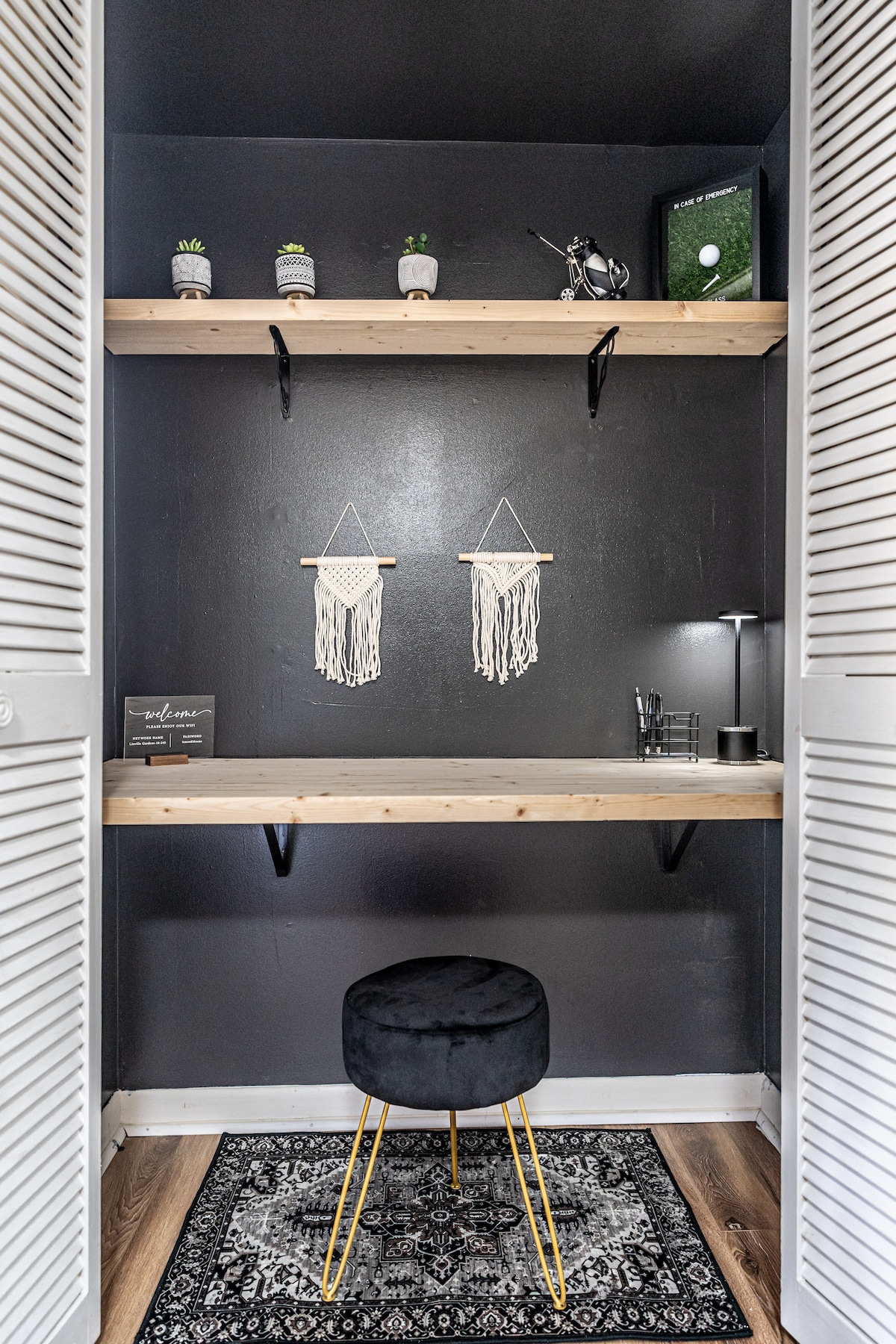 A small workspace is shown within a cozy nook, featuring a light wooden shelf supported by black brackets. A modern black stool with gold legs complements the tidy arrangement. Decorative plants and macramé wall hangings add a touch of character against the dark background.