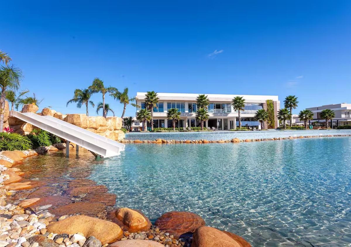 A tranquil swimming area features clear, turquoise waters with a natural rock slide extending into the pool. Palm trees are positioned around the landscape, complementing the serene environment. The modern building is visible in the background, enhancing the overall resort-like setting.