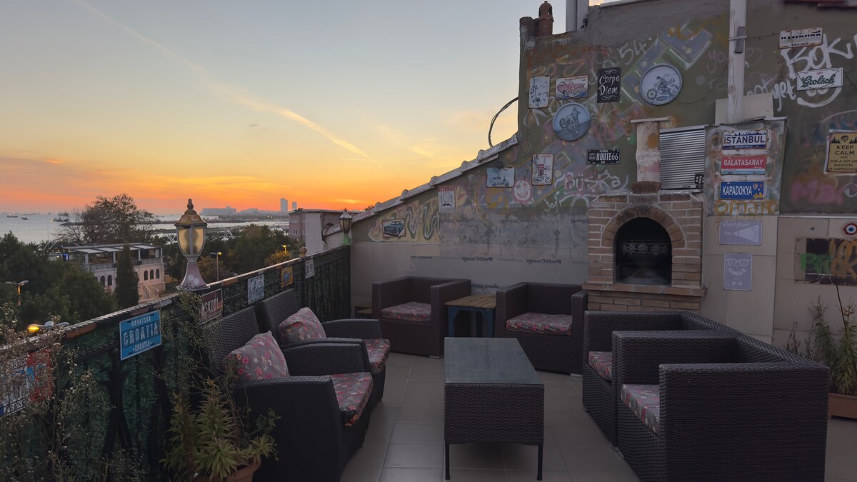 An outdoor terrace offers a view of a sunset over the skyline, surrounded by colorful murals and signs. Comfortable seating is provided, including a dark wicker seating set with cushions. A brick barbecue grill is positioned against a wall, completing the inviting atmosphere.
