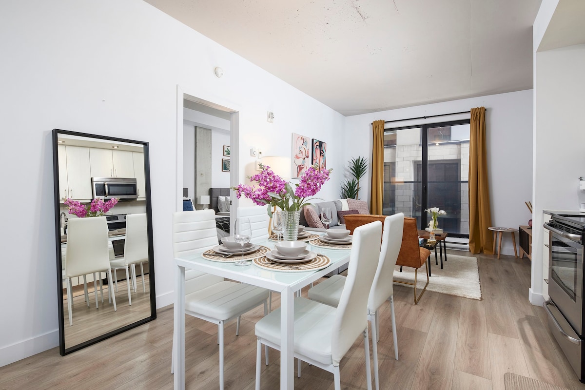 A bright dining area features a white table set with dishes and a floral centerpiece. Nearby, a living space is visible with a cozy couch and an accent chair, complemented by large windows dressed in gold curtains. A full-length mirror reflects the inviting atmosphere.