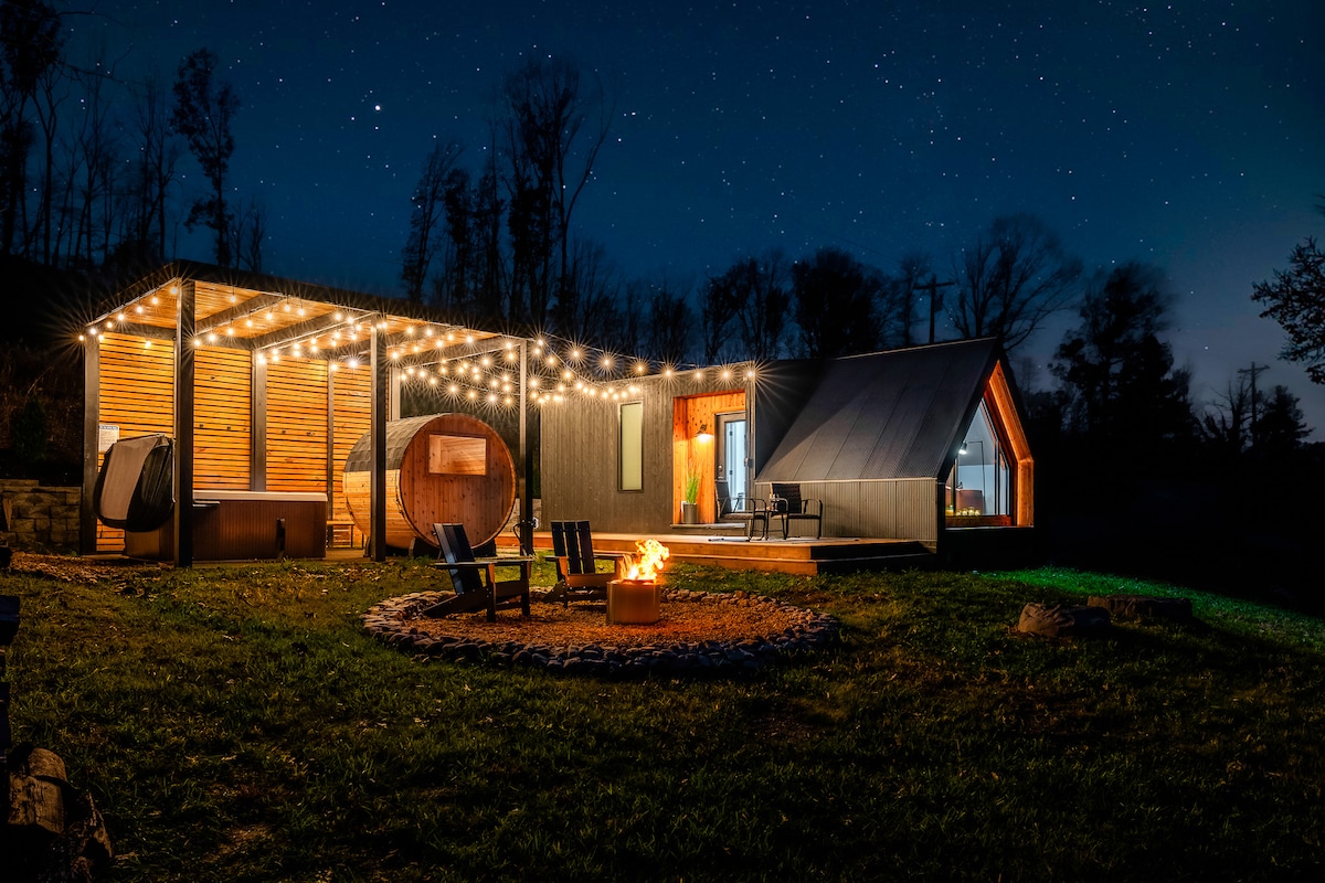 The cabin exterior is illuminated under a starry night sky, enhanced by string lights above the expansive deck. A cozy fire pit surrounded by chairs invites warmth. Nearby, the unique A-frame design stands against tall trees, creating a serene outdoor atmosphere.