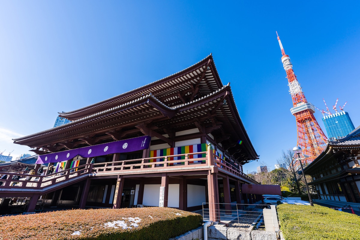 A traditional temple building is observed alongside the iconic Tokyo Tower, which stands tall against a clear blue sky. The temple features wooden architecture and colorful banners. A neatly maintained garden with manicured greenery surrounds the temple, providing a serene atmosphere.