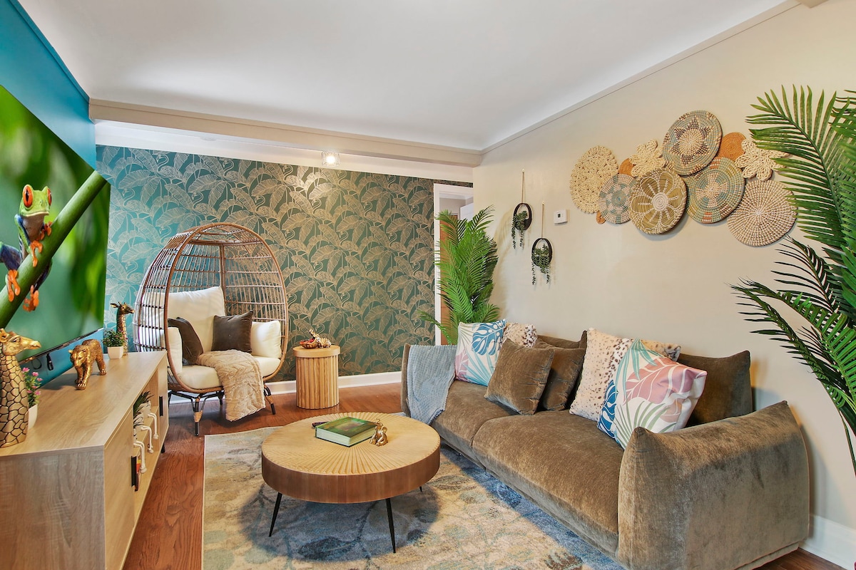 The living room features a cozy couch adorned with decorative pillows and a large circular coffee table at its center. A chic egg chair and vibrant wall art are complemented by tropical-inspired wallpaper and greenery, creating a welcoming and relaxed atmosphere.