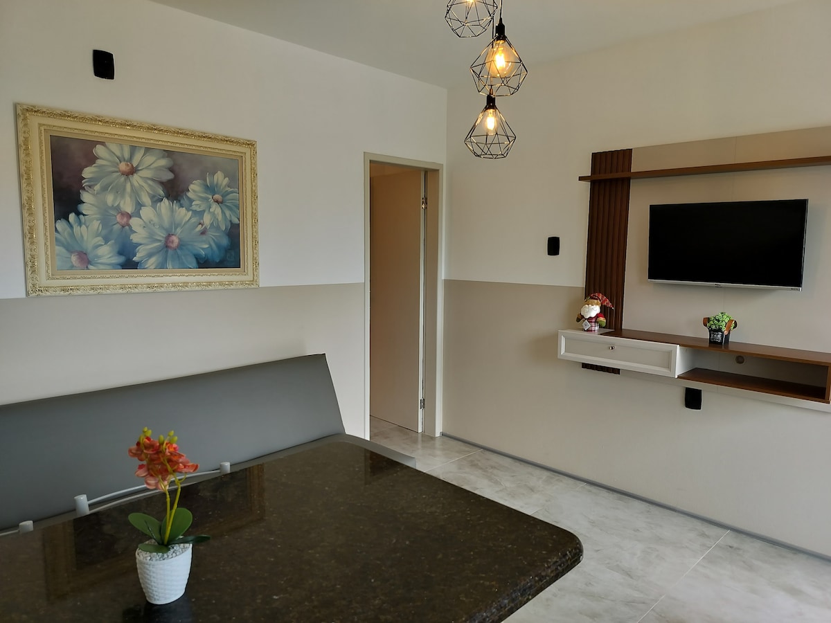 A casual dining area is highlighted with a dark stone table accented by a small potted plant. A large floral painting is displayed on the wall, while modern light fixtures hang from the ceiling. A mounted television is visible on the opposite wall.