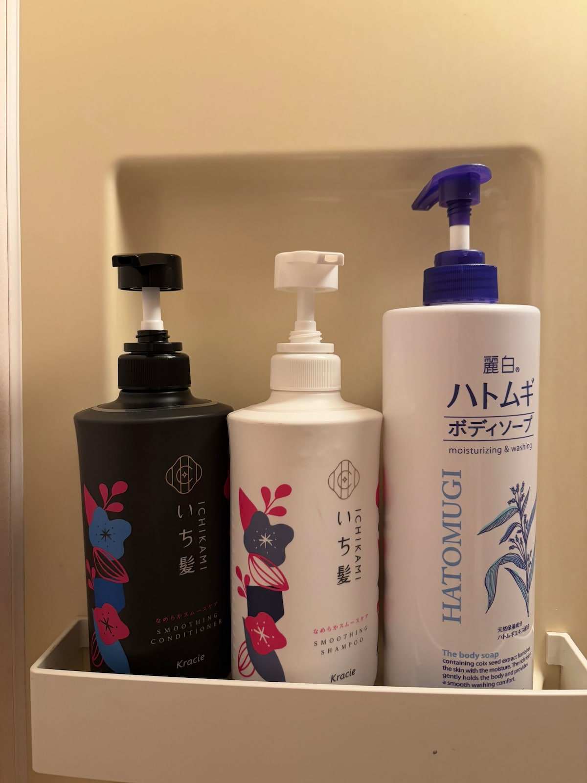 Three bottles of personal care products are displayed on a shelf. Each bottle features a pump dispenser, with varying colors and designs. The leftmost bottle has a dark floral design, while the center and right bottles are predominantly white, indicating different types of hair and body care.