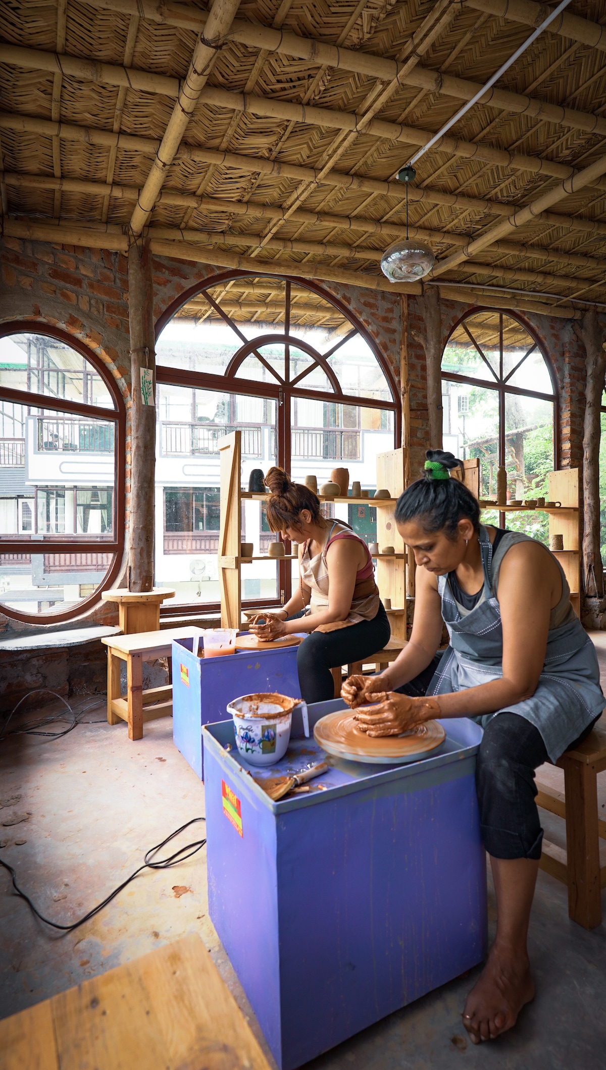 Two individuals are engaged in pottery creation within a bright workshop, surrounded by large windows framed in wood. They are seated at low tables with pottery wheels, showcasing a variety of tools and clay materials. Natural light enhances the warm, inviting atmosphere.