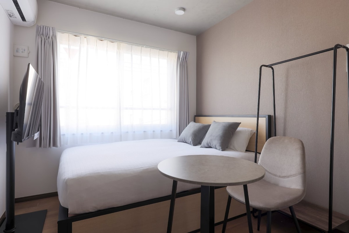 A cozy bedroom is presented with a spacious bed adorned with two pillows. Natural light filters through sheer curtains, illuminating the room. A small table and chair are positioned nearby, along with a wall-mounted monitor, creating a functional and welcoming space.