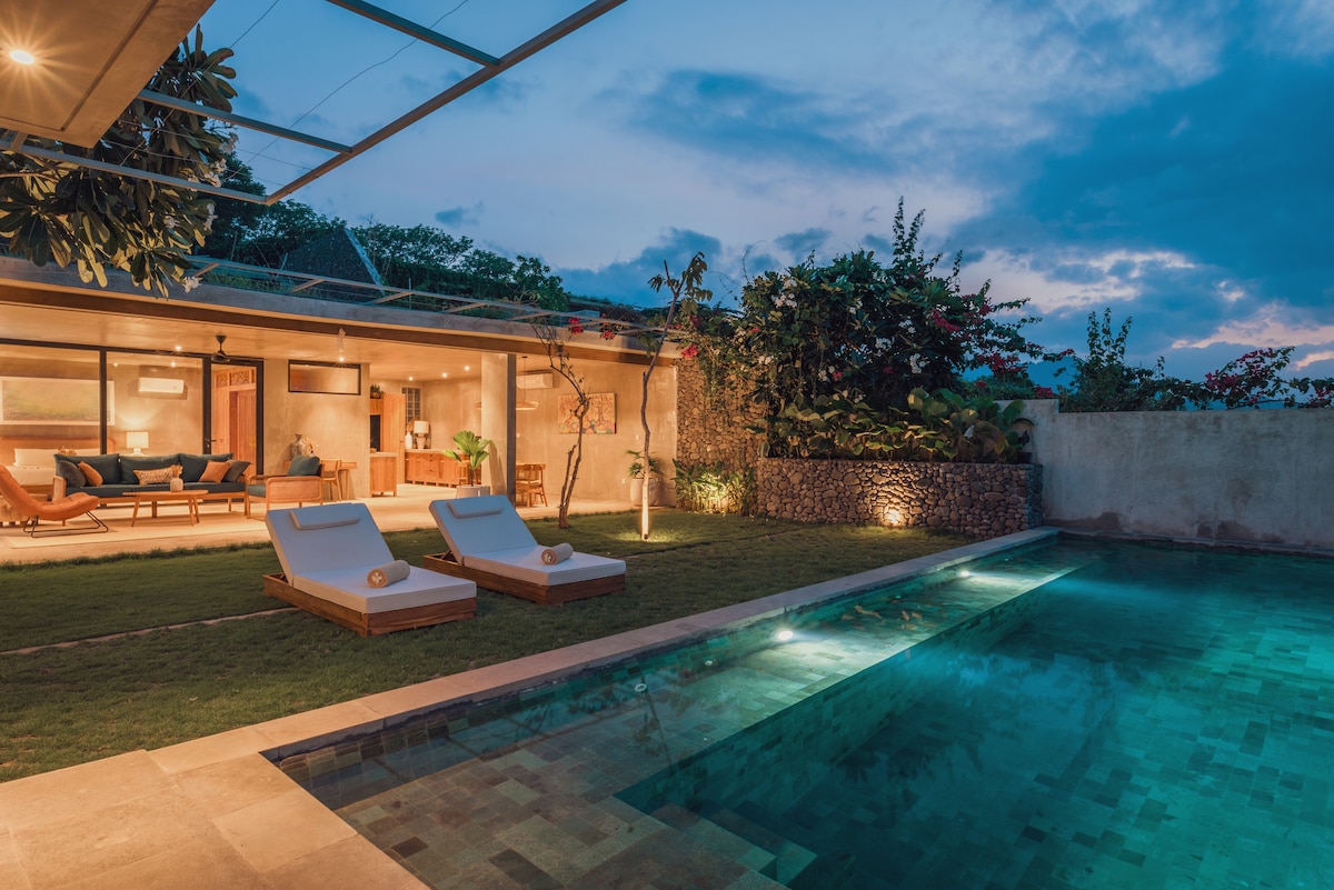 An outdoor area showcases a private pool accompanied by two lounge chairs. The pool's surface reflects the evening sky, while the surrounding garden is lush and dotted with flowering plants. Light glows softly from the villa's interior, creating a serene ambiance.