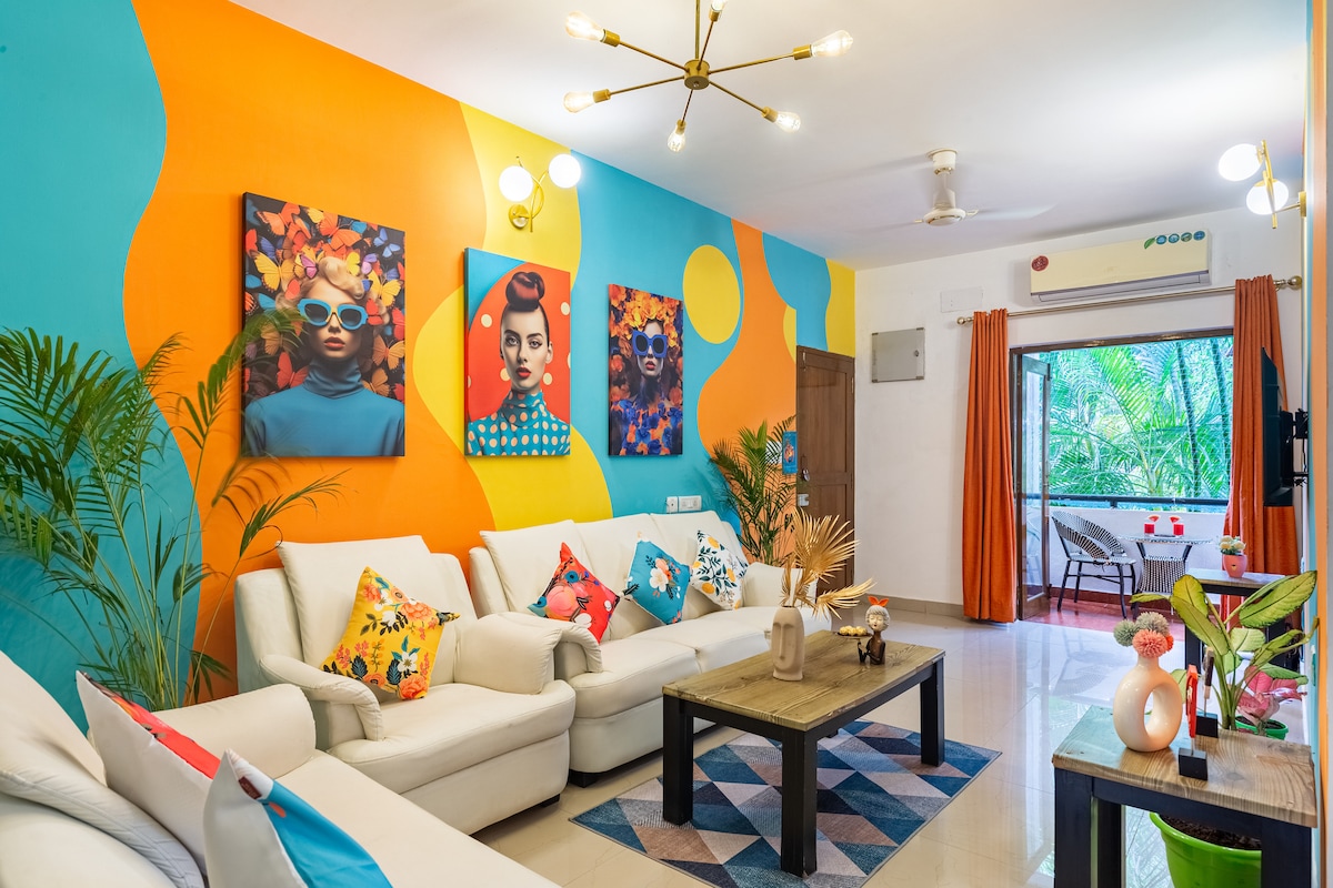 A vibrant living room featuring a colorful wall design with blue, orange, and yellow accents. The space contains a set of white sofas adorned with various patterned cushions. A wooden coffee table sits at the center, while large windows allow natural light to enhance the inviting atmosphere.