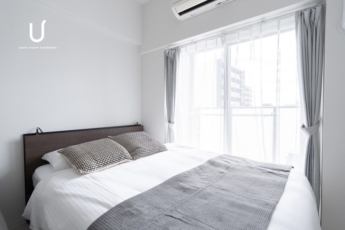 A bright sleeping area features a spacious bed with two textured pillows. Light filters through sheer curtains, illuminating the room. The walls are painted in a subtle tone that complements the overall design. One air conditioning unit is mounted near the window.