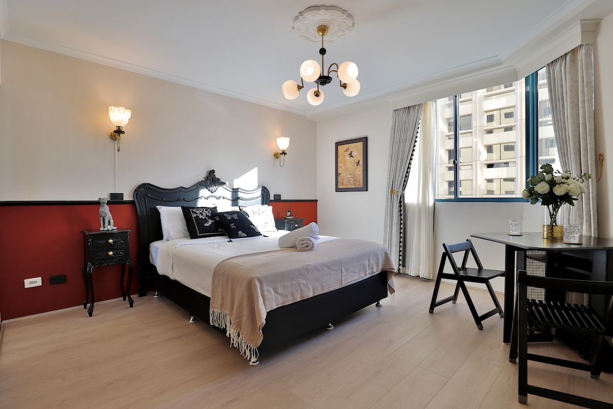 A stylish bedroom features a spacious bed adorned with white linens, complemented by a soft throw. Elegant lamps provide gentle illumination against a rich red accent wall. A small dining table with chairs is situated near the window, allowing natural light to fill the space.