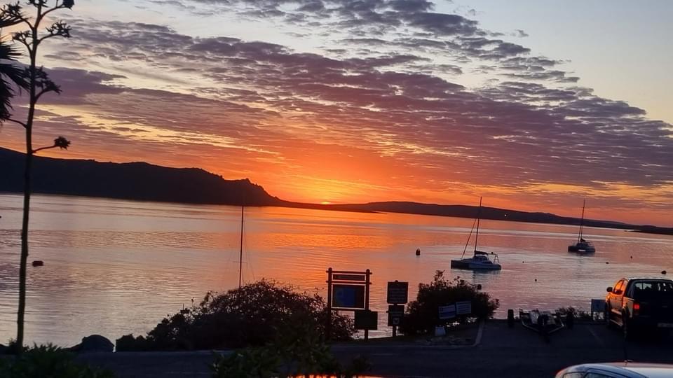 A serene sunset is captured over calm waters, with vibrant orange and purple hues filling the sky. Silhouetted boats are anchored in the foreground, while distant hills outline the horizon. The scene conveys a tranquil atmosphere perfect for relaxation.