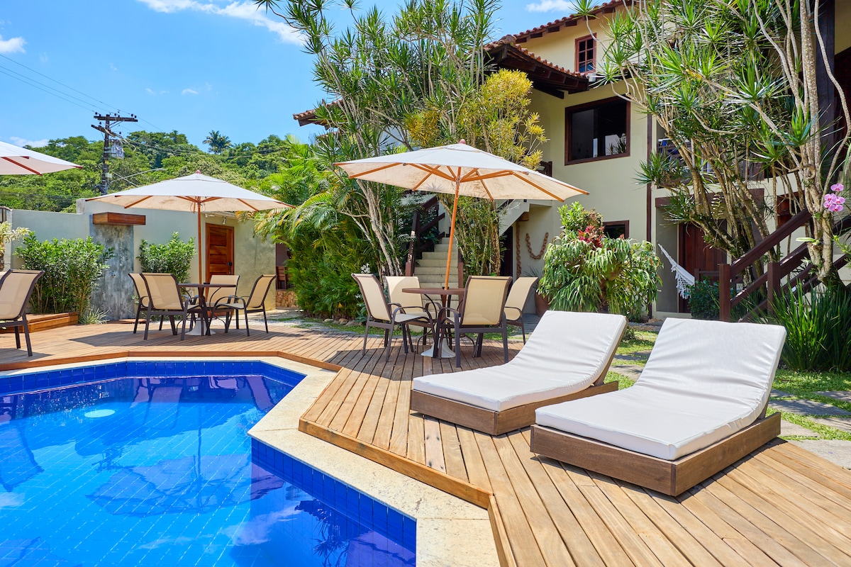 A serene outdoor area features a sparkling pool surrounded by comfortable lounge chairs and several shaded tables. Umbrellas provide refuge from the sun, and lush greenery adds to the calming atmosphere. The backdrop showcases a charming two-story house nestled among the trees.