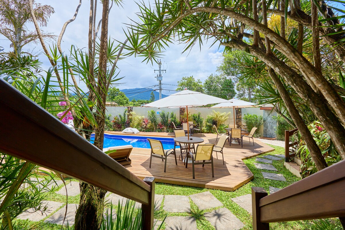 A well-maintained outdoor area features a swimming pool surrounded by lush greenery. Sun loungers and a circular dining table are positioned on a wooden deck, complemented by large umbrellas. The backdrop includes distant hills, contributing to the serene environment.