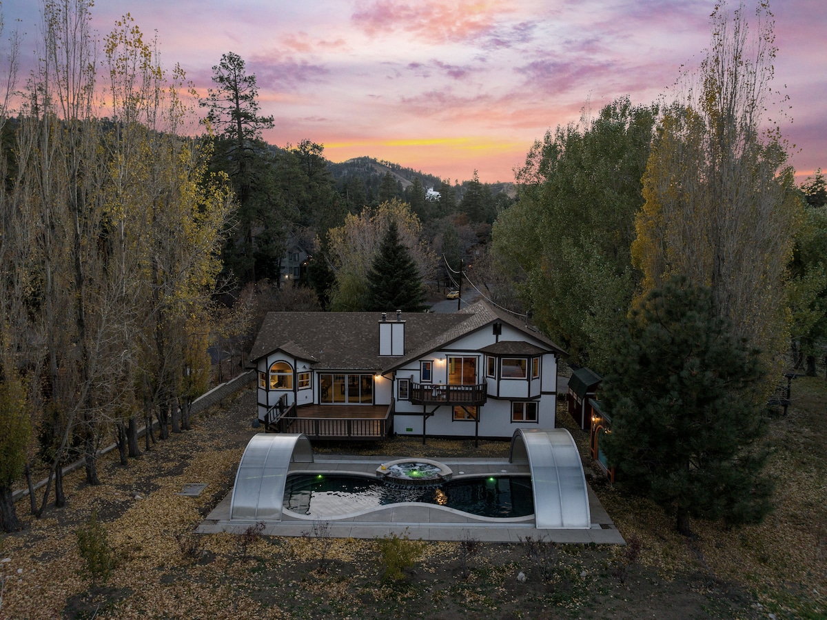 An aerial view of a cozy cabin framed by autumn foliage under a colorful sunset sky. The backyard features a heated pool beneath a protective cover, surrounded by landscaped grounds and trees, providing a serene outdoor setting.