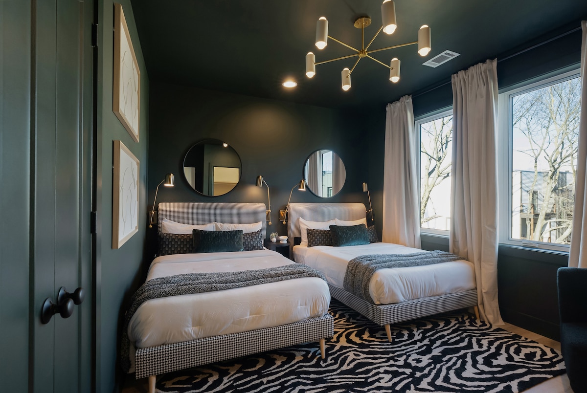 这间设计师卧室大胆时尚，配有两张带千鸟格纹布的床、斑马纹地毯、金色灯具和超大窗户，自然采光充足。
