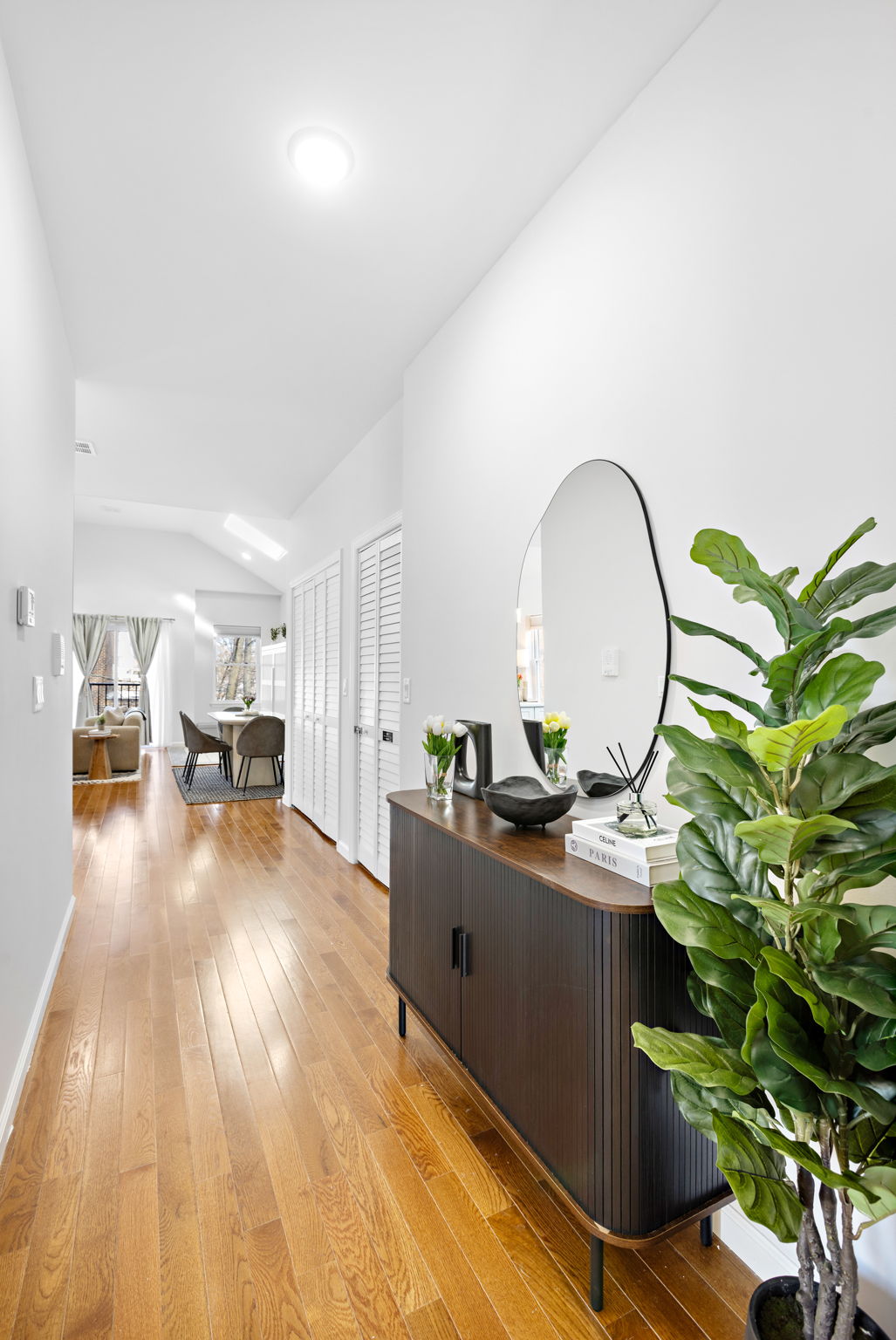 A bright hallway features polished hardwood floors and a modern console table with a circular mirror above. Greenery from a potted plant adds a pop of color, while distant views of a dining area are visible through open doors.