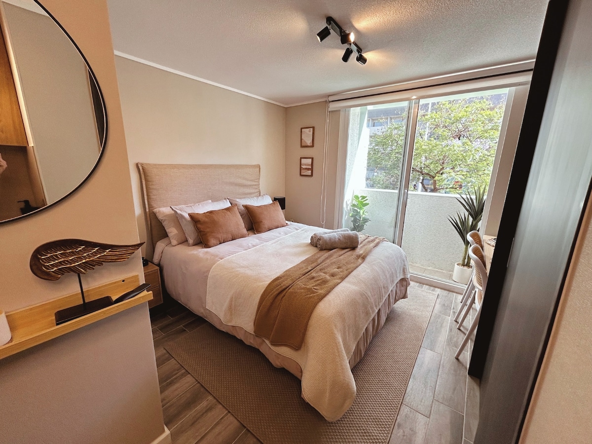 A bright bedroom features a spacious bed adorned with neutral-toned linens and decorative pillows. Natural light filters through glass doors leading to a small balcony, complementing the indoor plants. An inviting area rug and minimalist decor enhance the room's comfort.