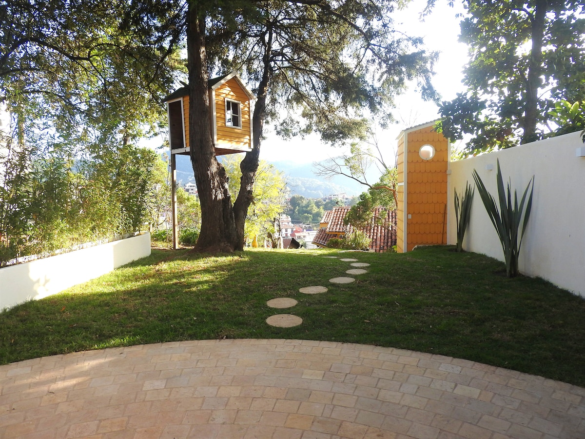 A well-maintained outdoor area features a grassy lawn and a pathway of stones leading to a charming treehouse. Sunlight filters through surrounding trees, enhancing the natural greenery. Views of the hillside and nearby rooftops are visible in the background, creating a peaceful setting.