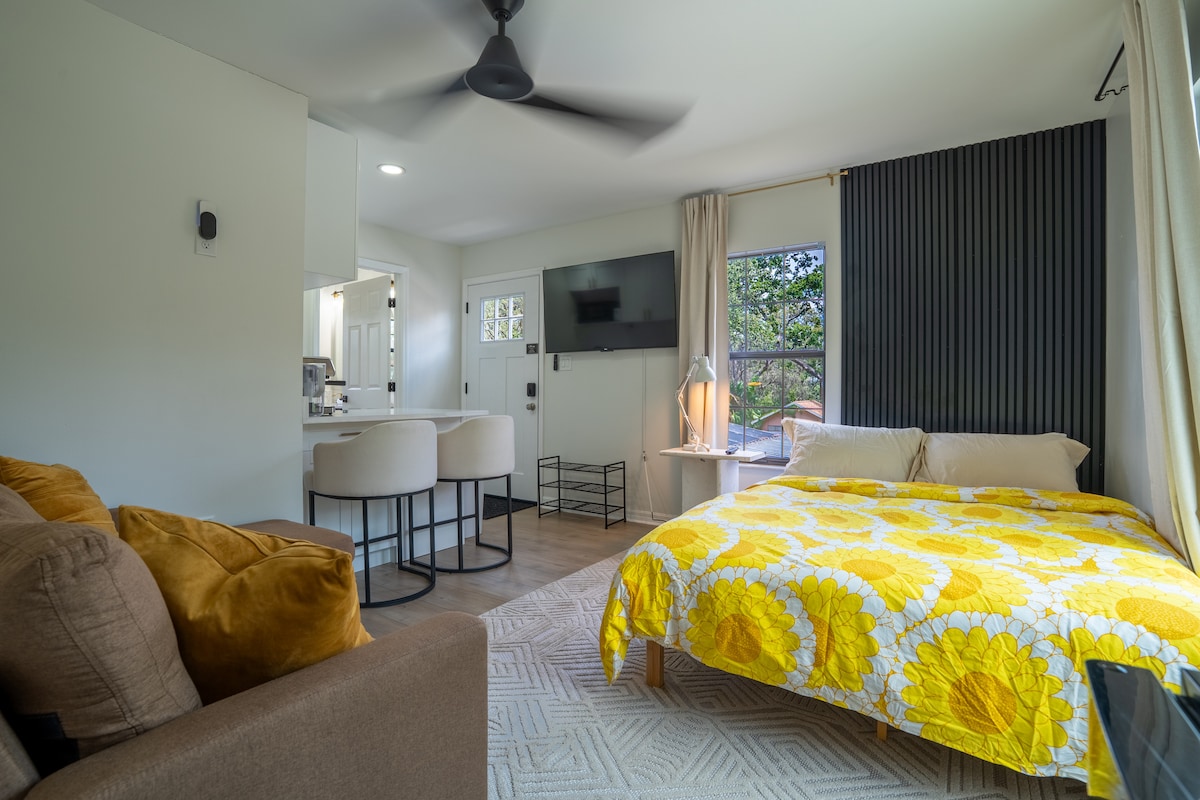 A bright studio suite features a spacious bed adorned with a vibrant yellow floral blanket. A comfortable seating area includes a brown sofa, while a light fixture and side table enhance the space. Large windows provide natural light, and a ceiling fan adds airflow.