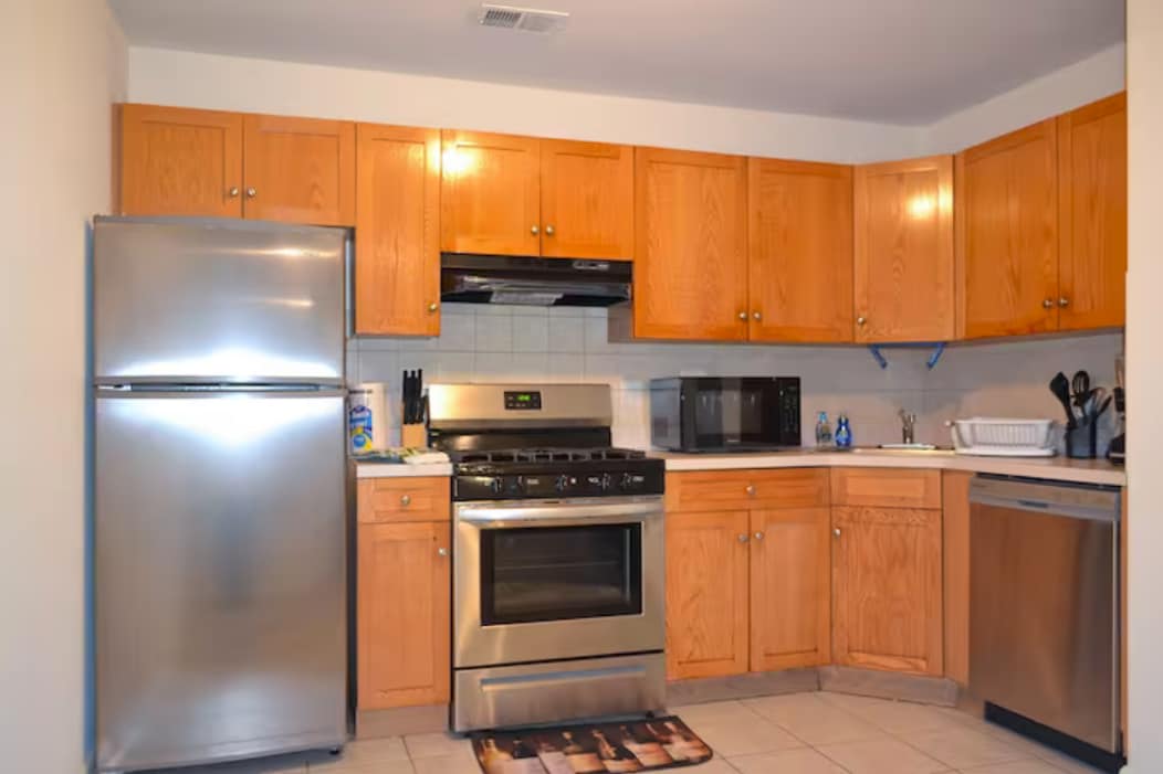 The kitchen features wooden cabinetry and stainless steel appliances, including a refrigerator and a stove. Counter space is maximized for food preparation, with essential utensils and a microwave within reach. A tiled floor enhances the practical design of the space.